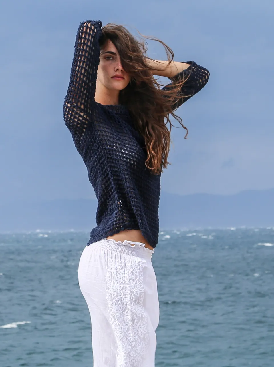 Femme en pull de crochet marine et pantalon blanc brodé, cheveux au vent devant la mer à Porquerolles.