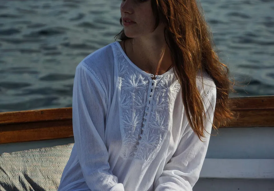 Détail d'une tunique blanche à broderie de feuilles, femme assise dans un bateau en bois à Porquerolles, lumière dorée.