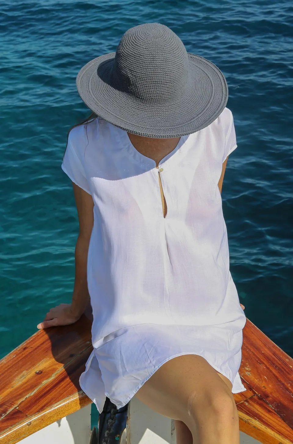 Femme de dos sur l'étrave d'un pointu, robe blanche et capeline crochetée grise au-dessus de l'eau à Porquerolles.