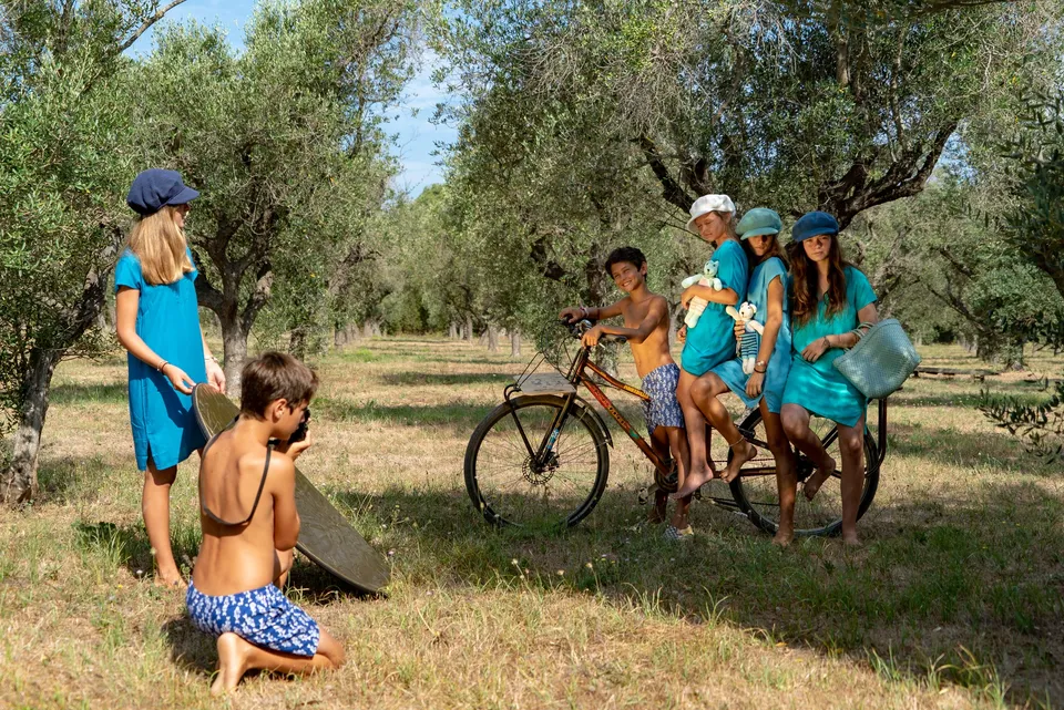 Enfants en robes-tuniques turquoise et casquettes autour d'un vieux vélo dans une oliveraie ensoleillée à Porquerolles.