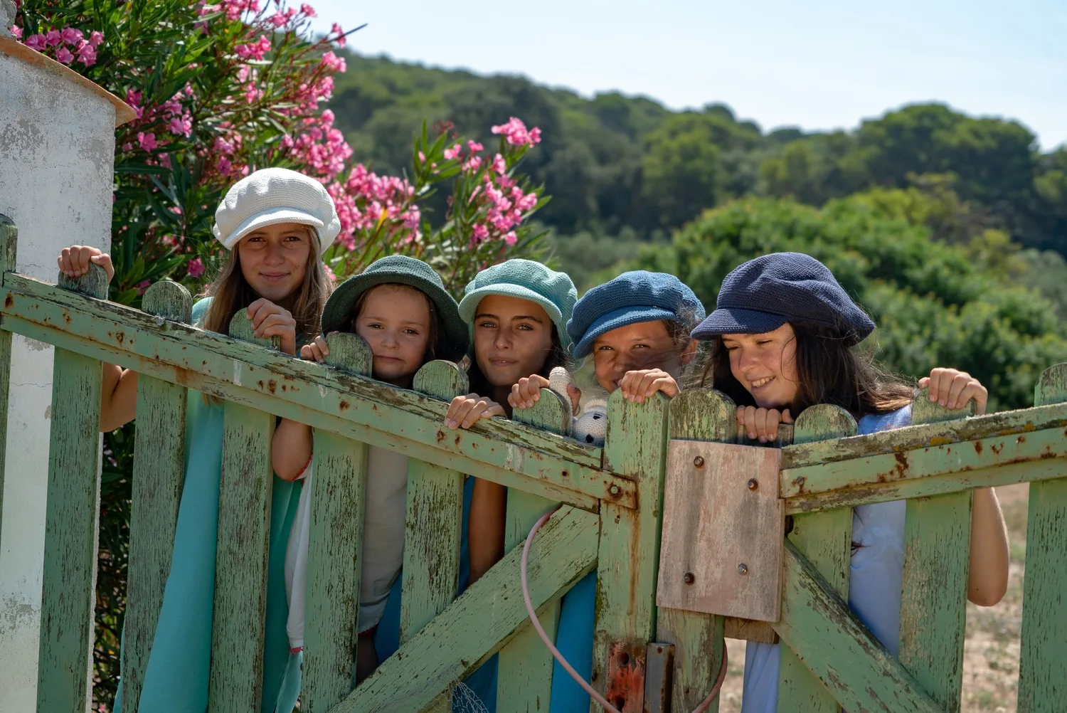 Cinq enfants en casquettes et bobs de crochet colorés, penchées sur un portail de bois sous les lauriers-roses à Porquerolles.