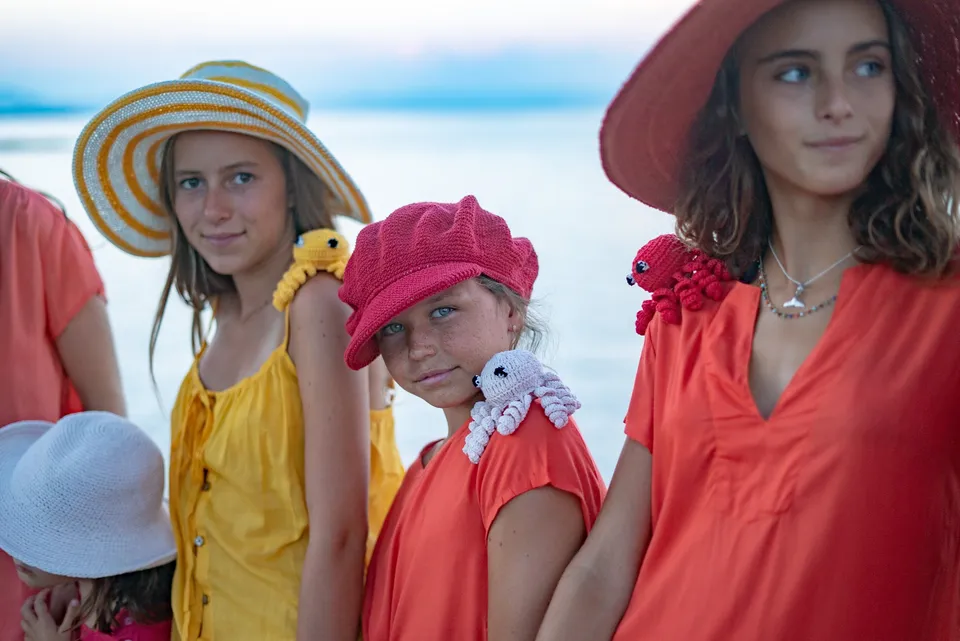 Jeunes filles en tuniques corail et jaune, chapeaux et pieuvres crochetées devant la mer pâle à Porquerolles.