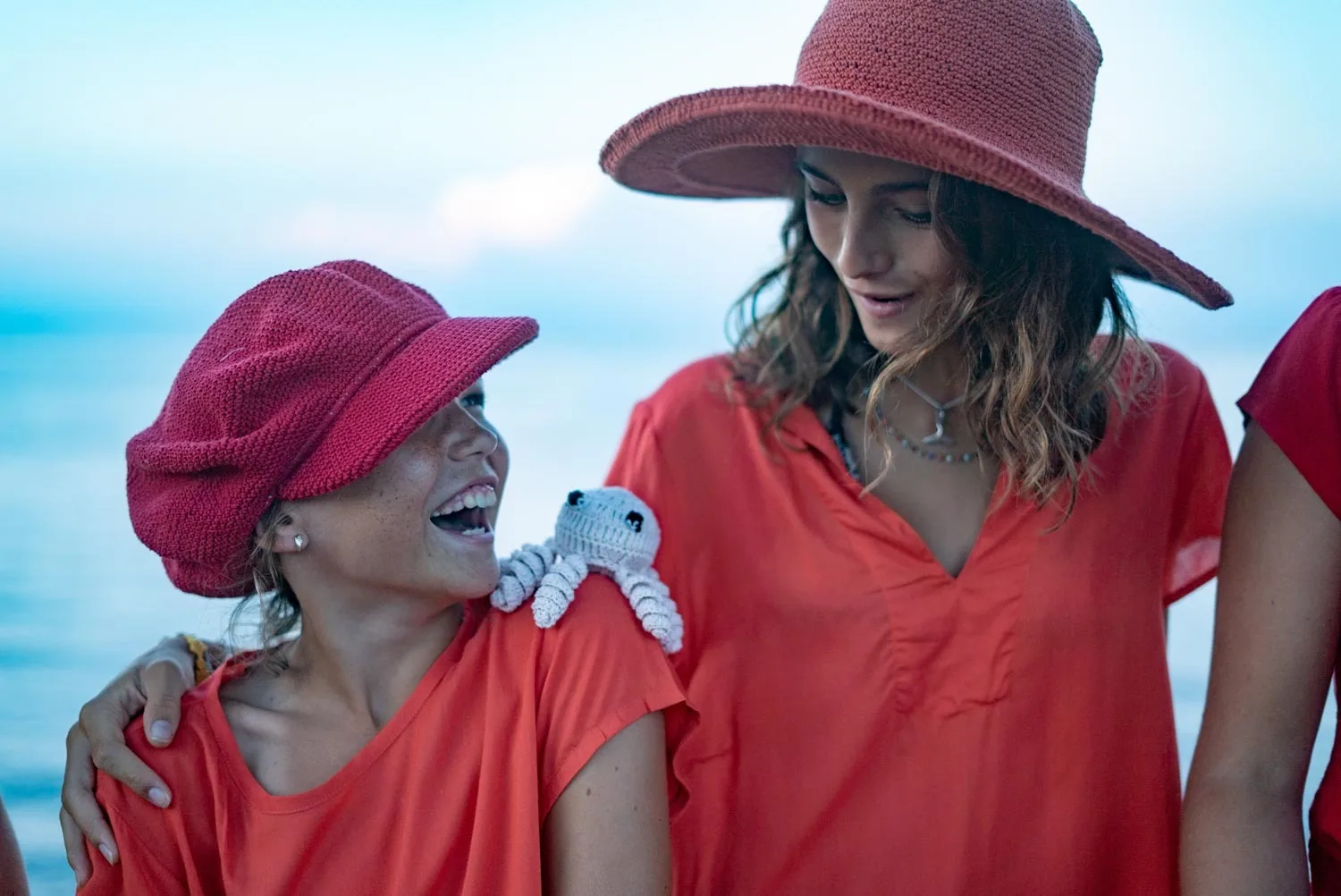 Deux jeunes filles en tuniques corail et chapeaux de crochet rient devant la mer au crépuscule à Porquerolles.
