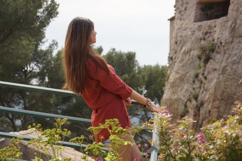 Femme en tunique terracotta appuyée à une rampe verte devant un mur de pierre et la verdure à Porquerolles.
