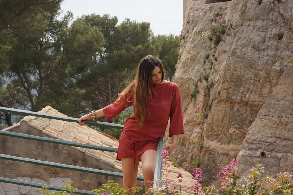 Femme en tunique terracotta appuyée à une rampe verte, mur de pierre et toits à Porquerolles.