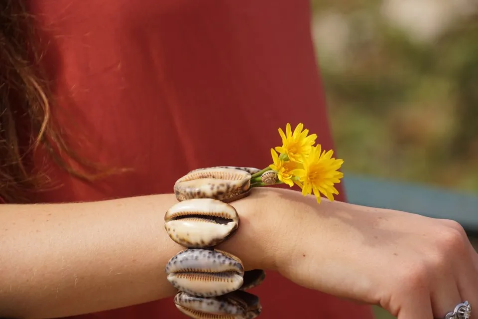 Bracelet de coquillages cauris au poignet, petites fleurs jaunes glissées dessous, robe brique, à Porquerolles.