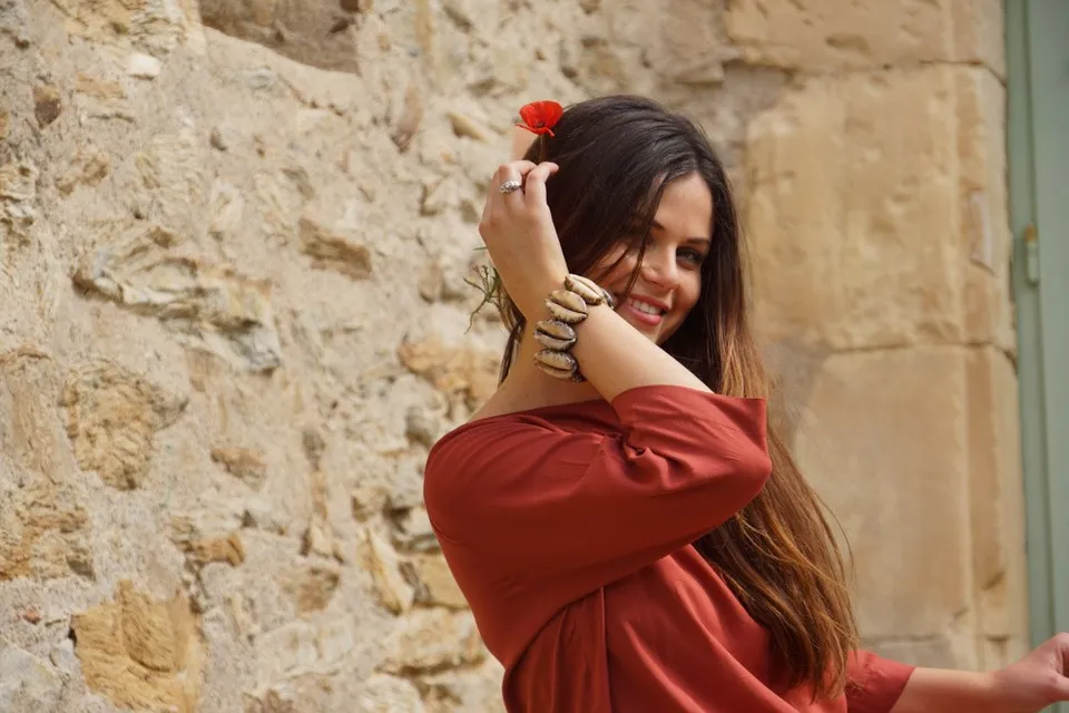 Femme souriante en tunique terre cuite, fleur rouge dans les cheveux, devant un mur de pierres à Porquerolles.