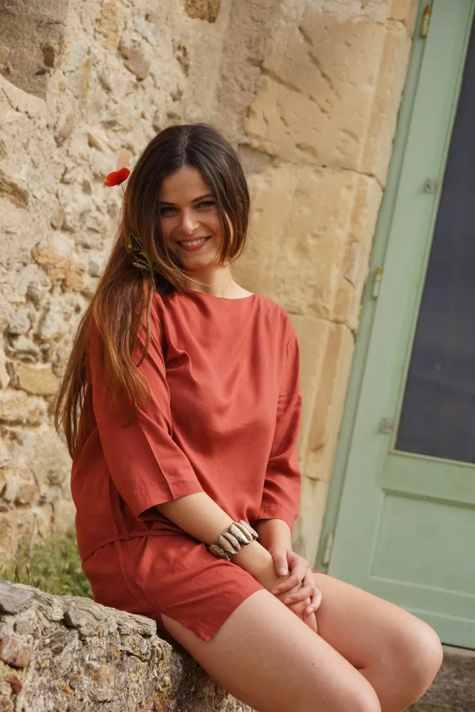 Femme en tunique terracotta assise contre un mur de pierre et une porte verte, bracelet au poignet à Porquerolles.