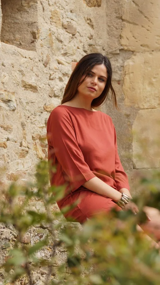 Femme en tunique terracotta devant un mur de pierres anciennes dans une ruelle de Porquerolles.