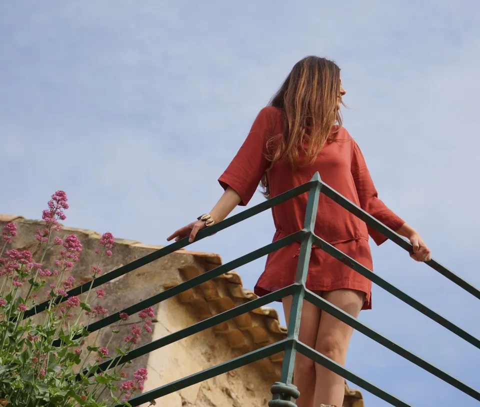 Femme en tunique terracotta derrière une rambarde verte, fleurs roses et toit de tuiles sous ciel pâle à Porquerolles.