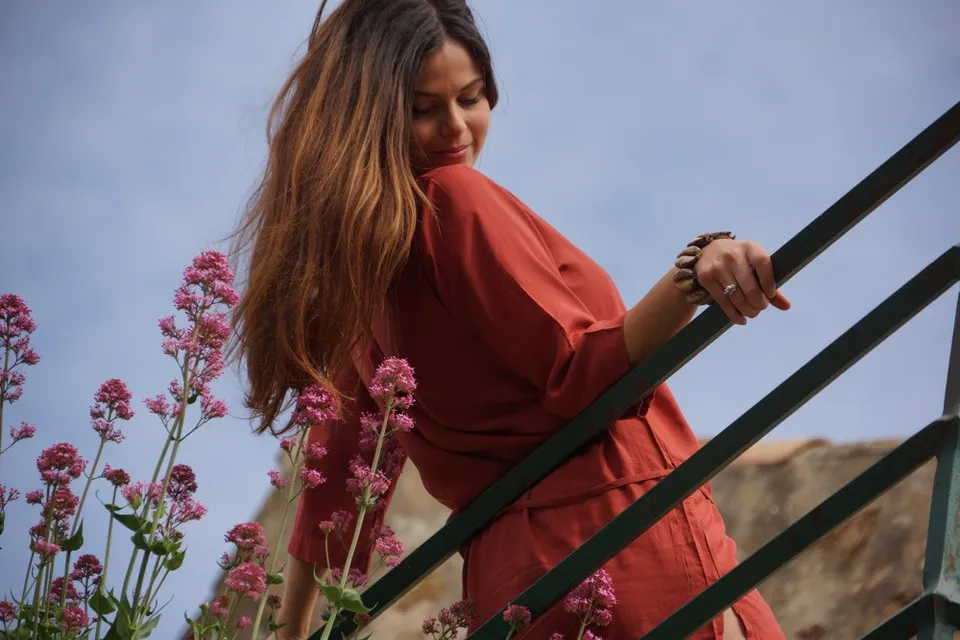 Femme en tunique terracotta près de fleurs roses sur un escalier à rambarde verte dans le village de Porquerolles.