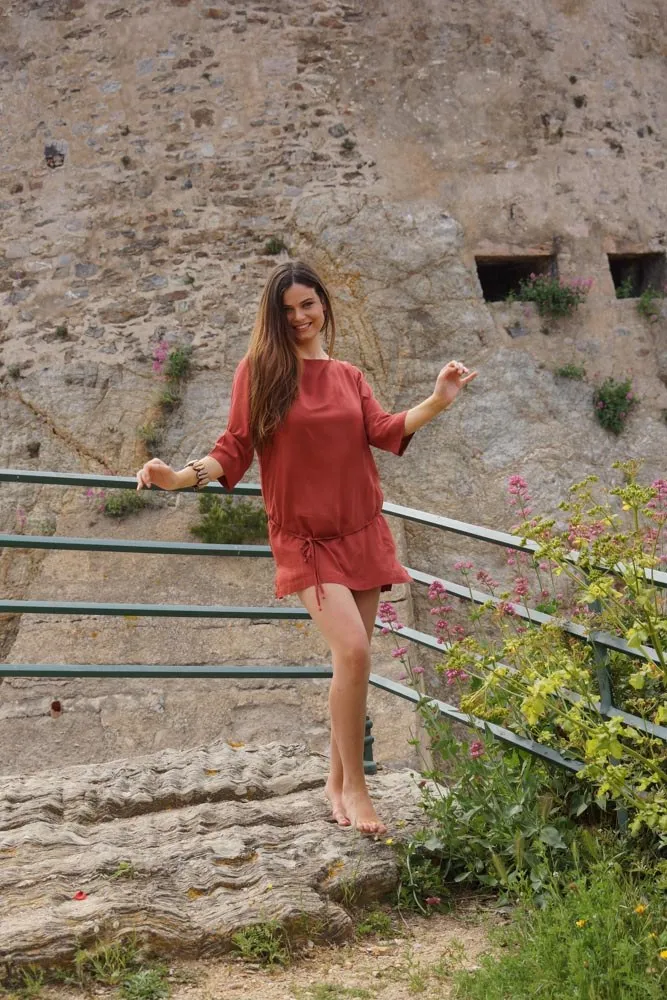Femme en tunique terracotta debout pieds nus devant un rempart de pierre à Porquerolles.