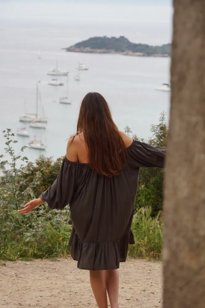 Femme de dos en robe grise fluide contemplant une baie de voiliers sous ciel couvert à Porquerolles.