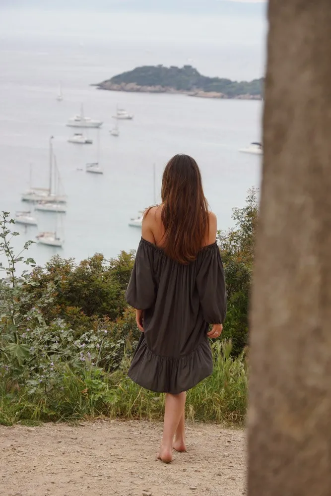 Femme de dos pieds nus en robe noire épaules nues face à la baie et aux bateaux ancrés à Porquerolles.