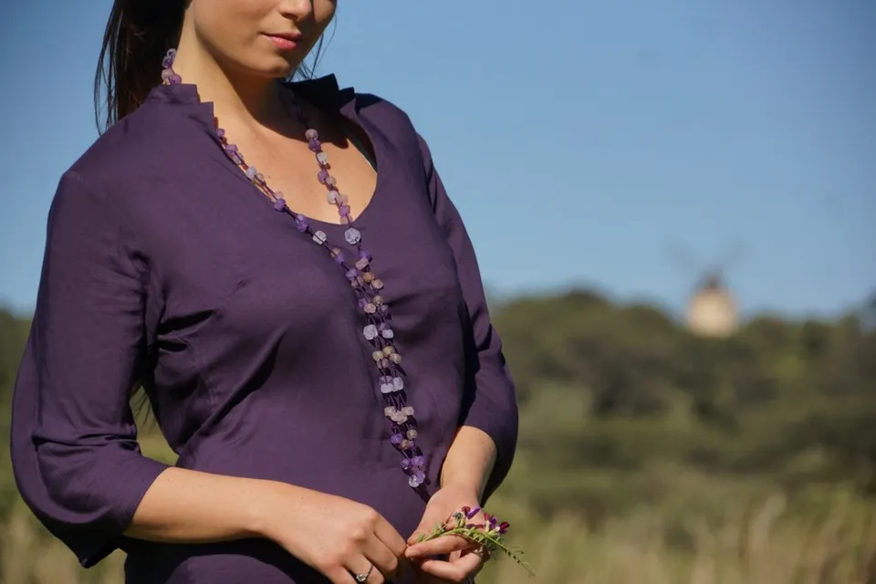 Femme en tunique mauve et collier de perles, tenant une petite fleur mauve, maquis et moulin flous à Porquerolles.