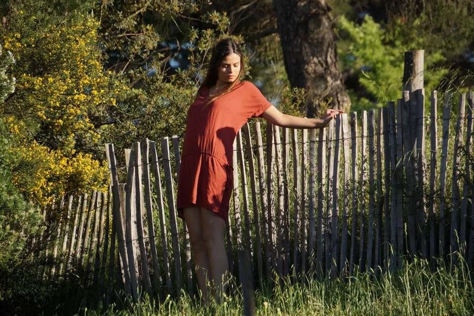 Femme en robe terracotta appuyée à une clôture de bois dans le maquis doré de fin de journée à Porquerolles.