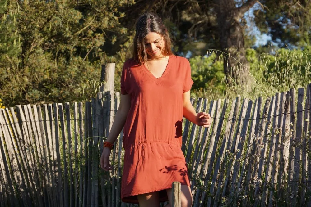 Femme en robe terracotta souriante, bracelet perlé, devant une ganivelle et le maquis doré à Porquerolles.
