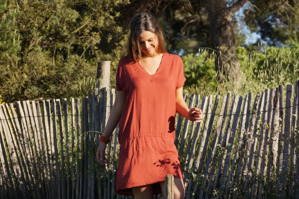 Femme en robe terracotta souriante, bracelet perlé, devant une ganivelle et le maquis doré à Porquerolles.