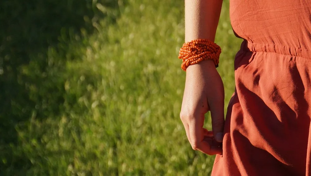 Main le long d'une robe terracotta, bracelet de perles orange au poignet, herbes au soleil, à Porquerolles.