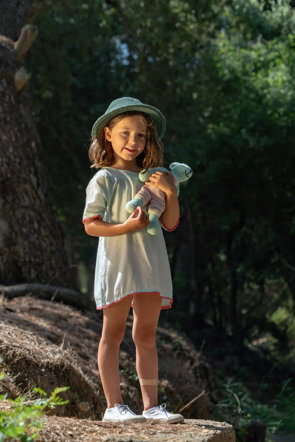 Fillette souriante en robe menthe ourlée de rouge, chapeau de crochet vert et ours en crochet sous les pins à Porquerolles.