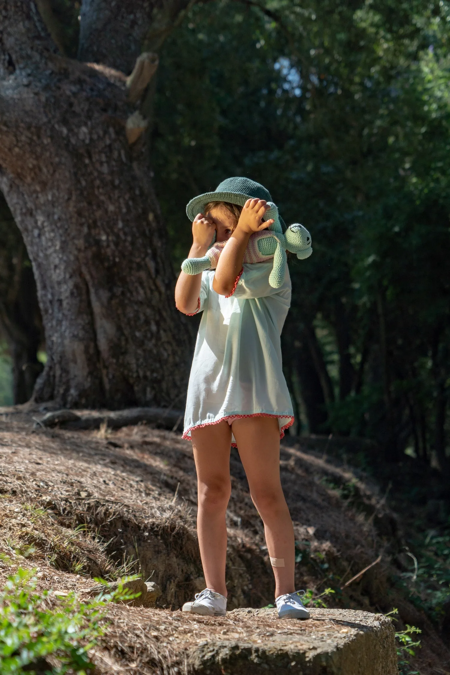 Enfant en tunique claire et chapeau vert tenant une tortue au crochet sous un arbre du maquis de Porquerolles.