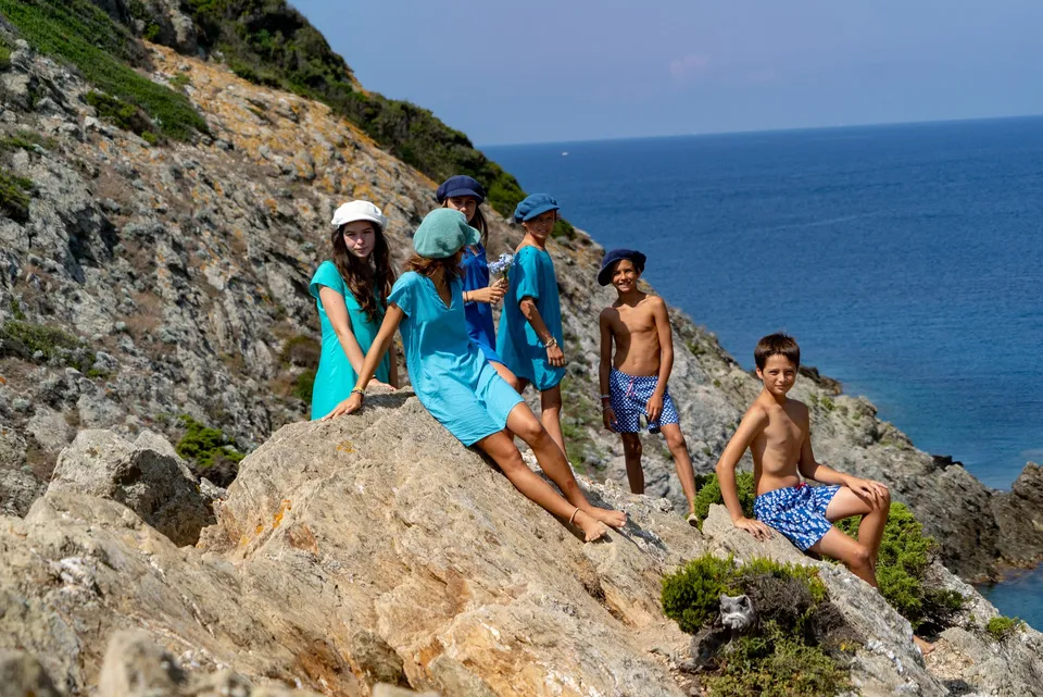 Six enfants en turquoise et bleu, shorts imprimés et chapeaux, sur des rochers face à la mer de Porquerolles.