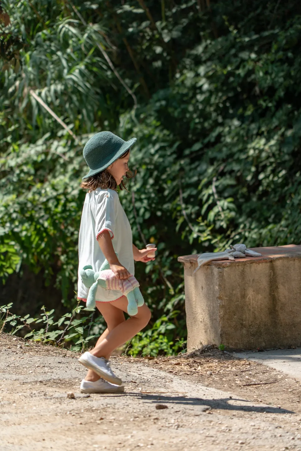 Petite fille en tunique claire ourlée de rose et chapeau vert, doudou sous le bras sur un sentier à Porquerolles.