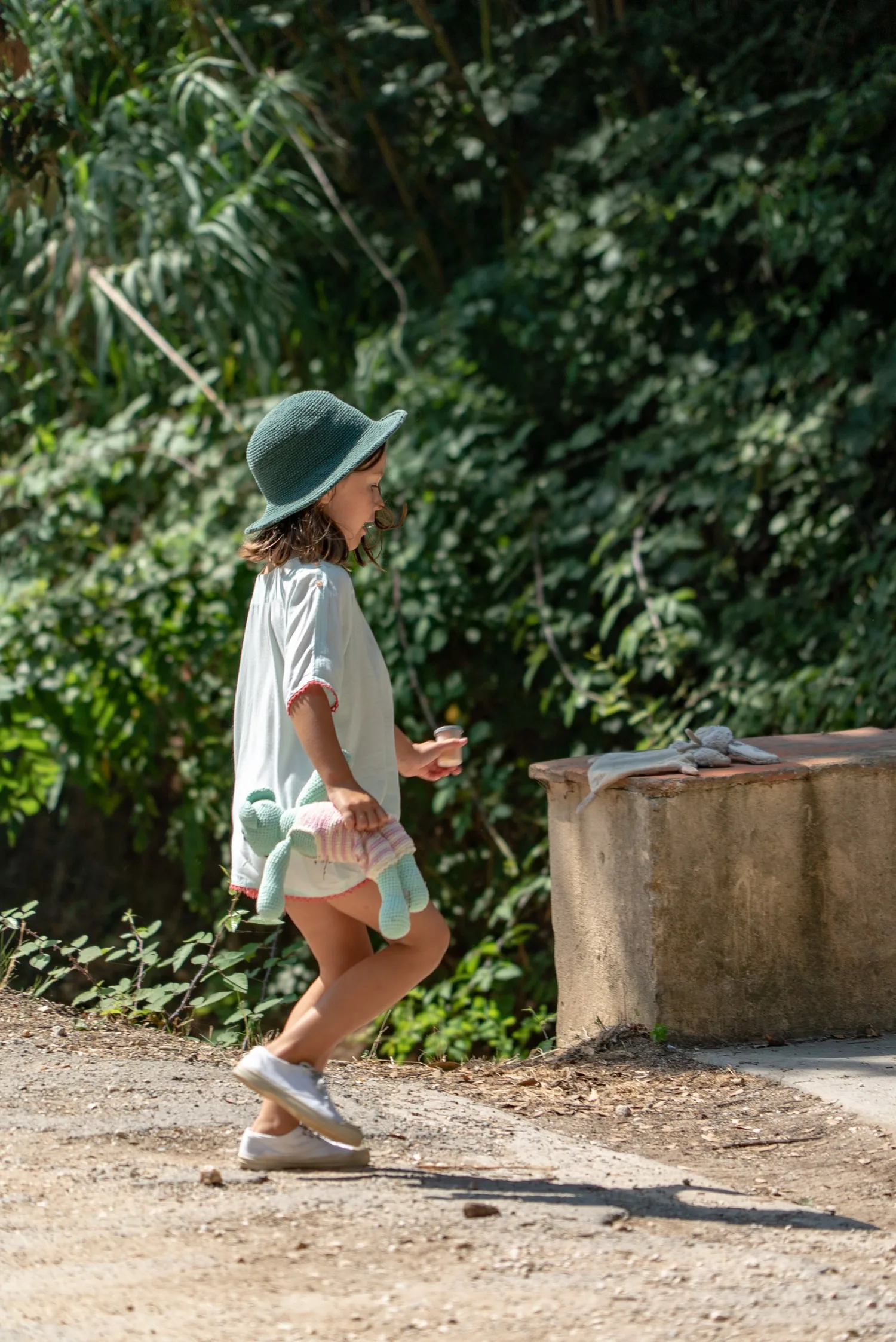 Petite fille en tunique claire ourlée de rose et chapeau vert, doudou sous le bras sur un sentier à Porquerolles.