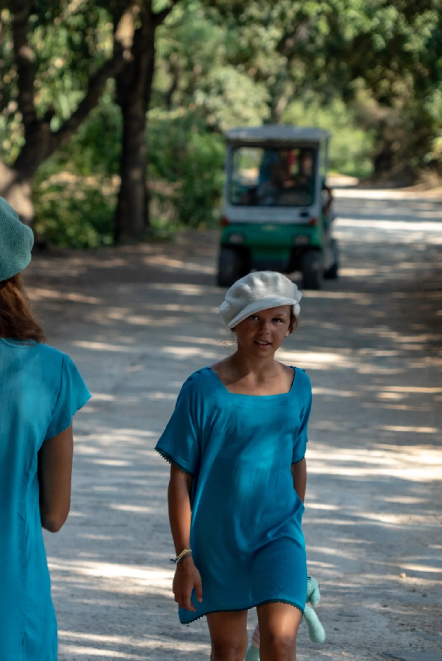 Fillette en tunique turquoise et casquette blanche marchant sur un chemin ombragé du maquis à Porquerolles.