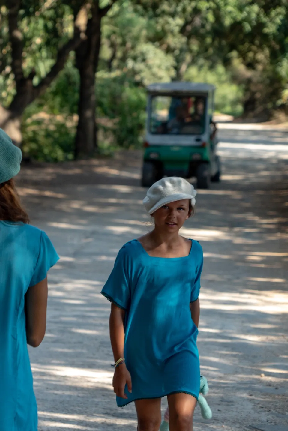 Fillette en tunique turquoise et casquette blanche marchant sur un chemin ombragé du maquis à Porquerolles.