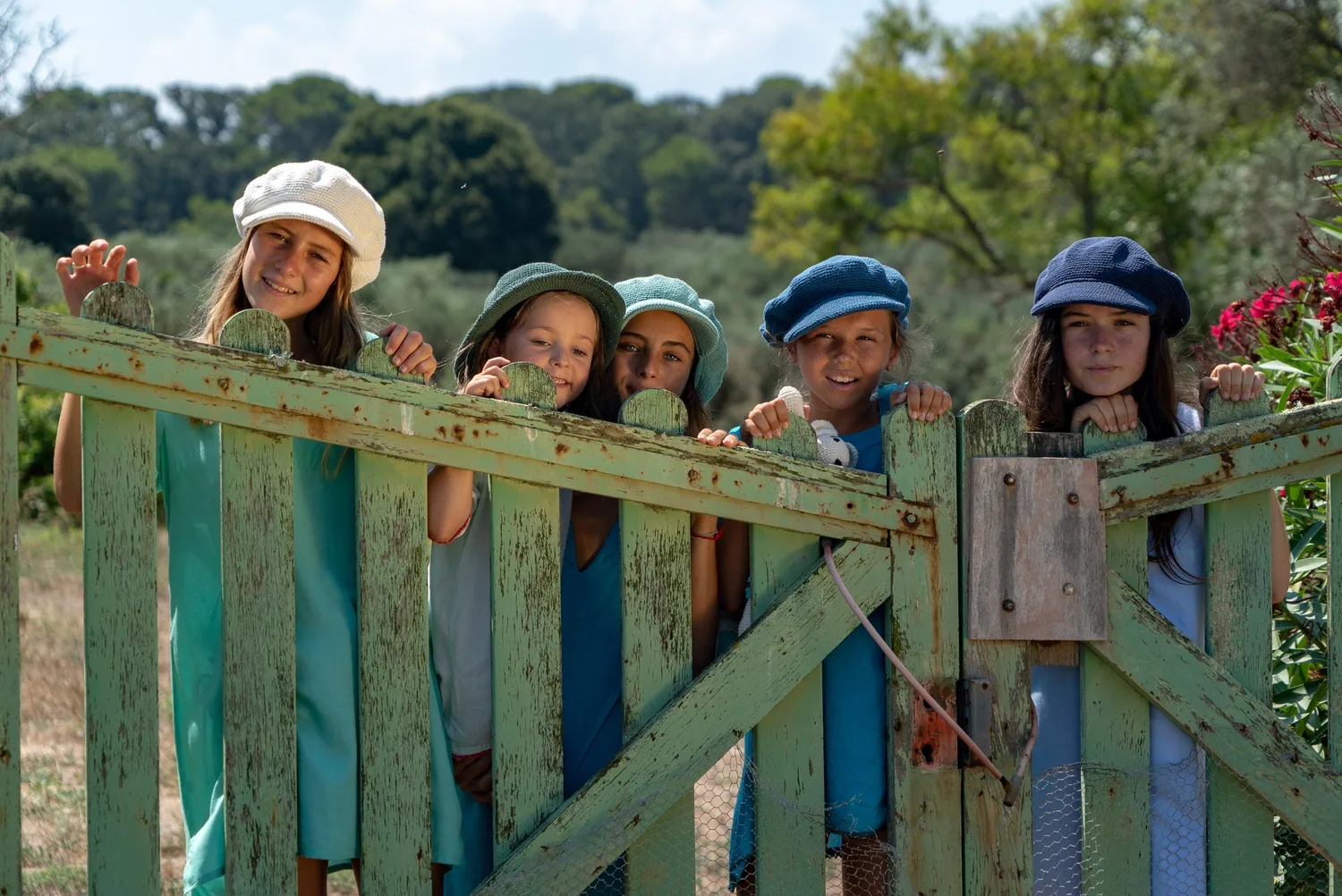 Cinq enfants derrière une clôture verte, casquettes colorées, oliviers et fleurs roses à Porquerolles.