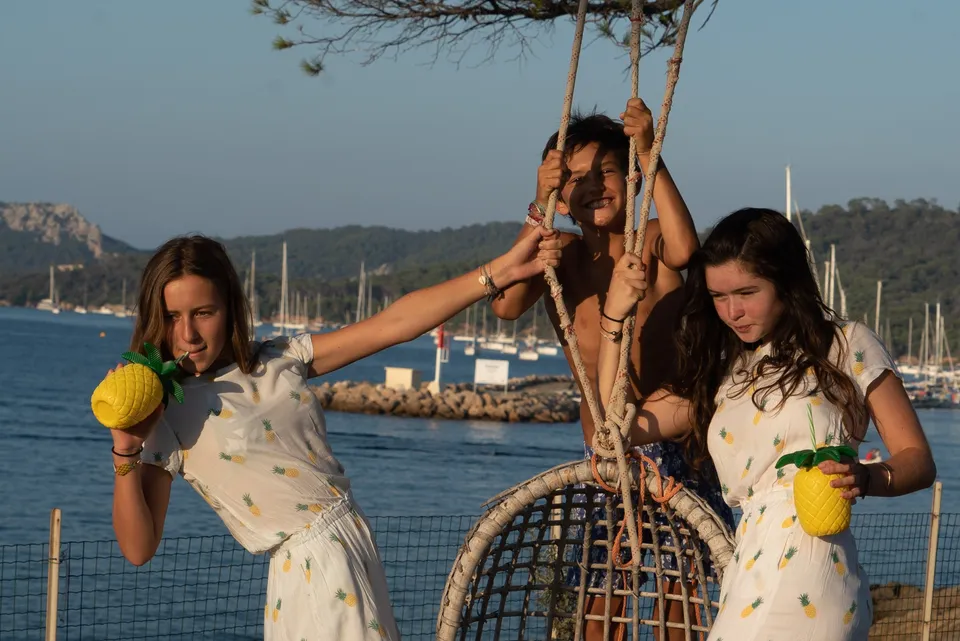 Enfants en robes blanches à motifs ananas près d'une chaise en vannerie, voiliers ancrés derrière à Porquerolles.