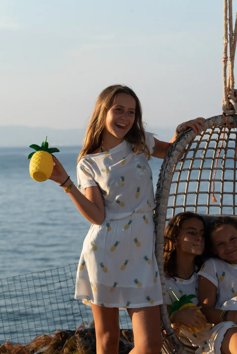 Trois jeunes filles en robes blanches à ananas, fauteuil œuf en vannerie face à la mer dorée à Porquerolles.
