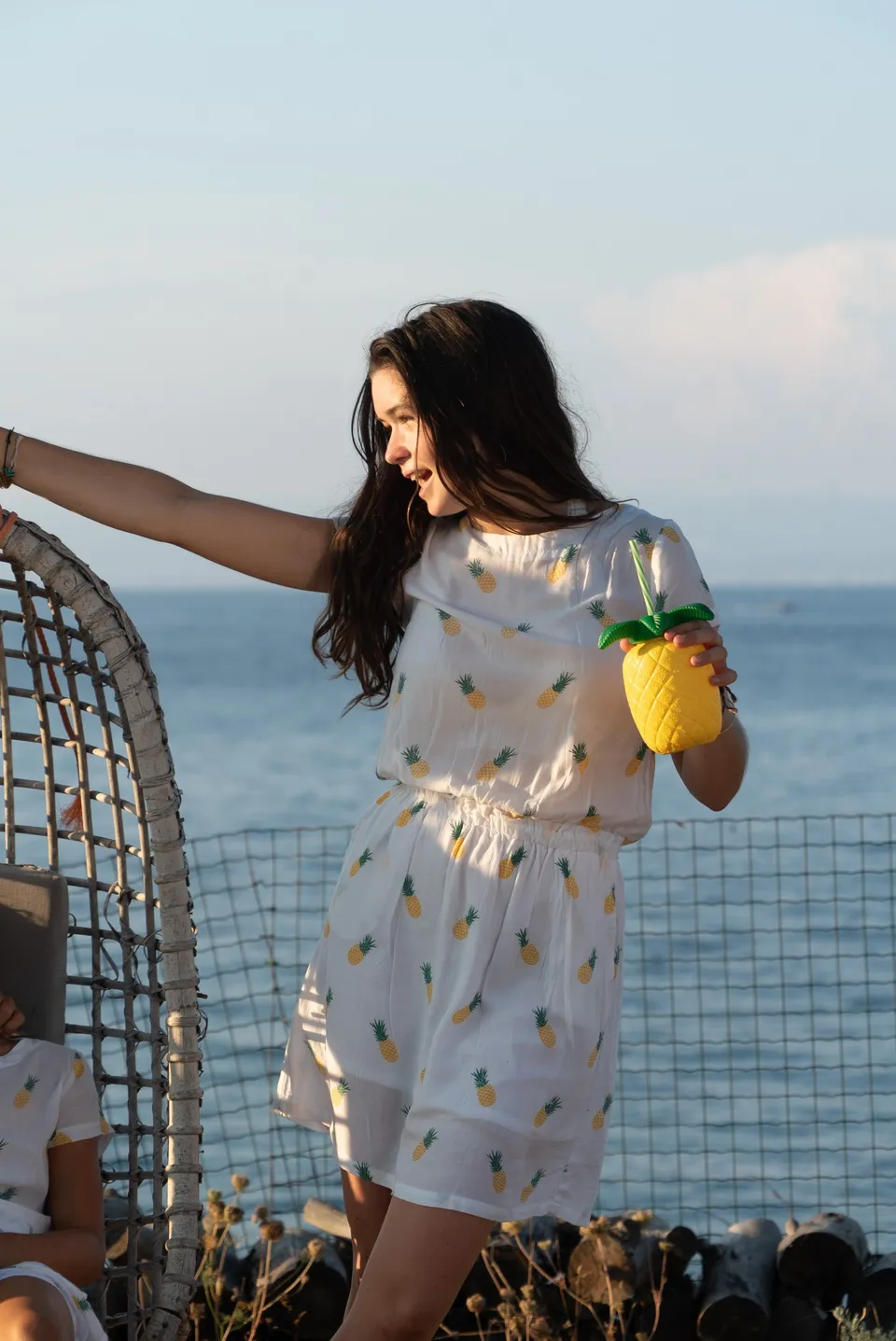 Jeune fille en robe blanche à ananas tenant un gobelet ananas sur une terrasse face à la mer à Porquerolles.