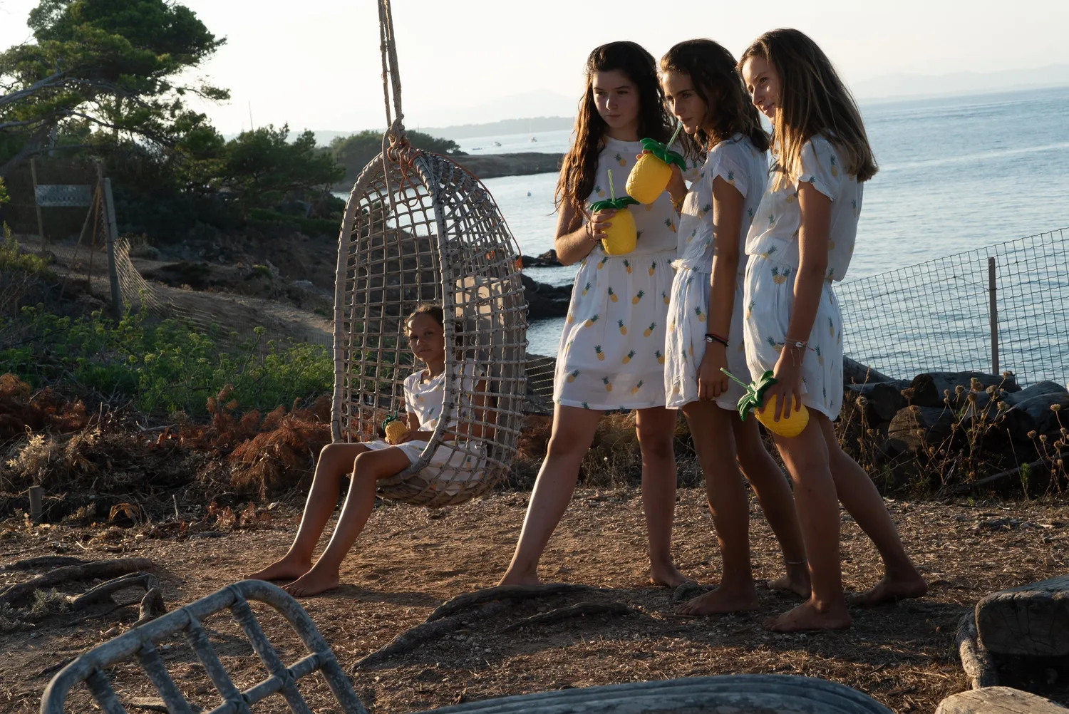 Filles en robes blanches à motifs ananas autour d'un fauteuil en vannerie face à la mer à Porquerolles.