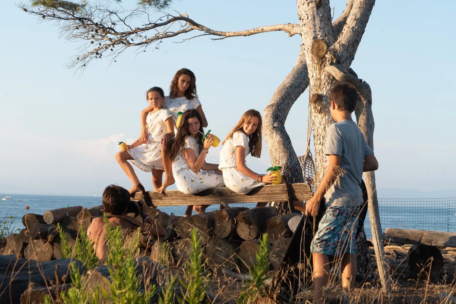 Quatre filles en robes à motifs ananas posées sur des rondins face à la mer au crépuscule à Porquerolles.