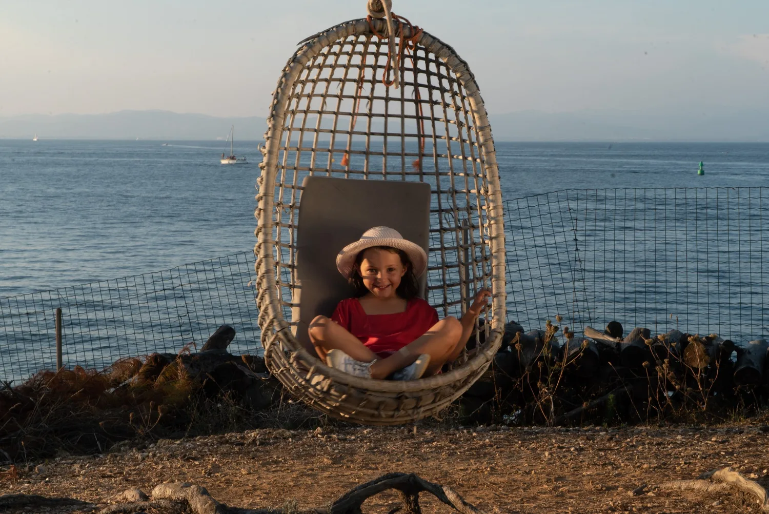Fille en tunique rouge et chapeau clair dans une chaise d'osier suspendue face à la mer de Porquerolles.