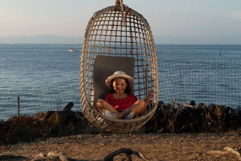 Fille en tunique rouge et chapeau clair dans une chaise d'osier suspendue face à la mer de Porquerolles.