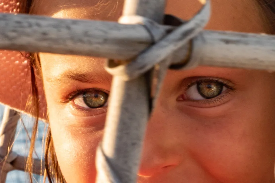 Yeux verts d'un enfant vus entre les lattes d'une barrière blanche patinée, lumière basse, à Porquerolles.