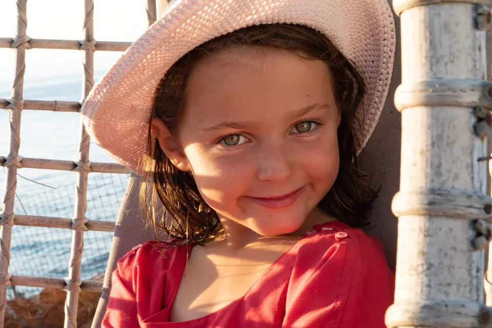 Petite fille en bonnet au crochet rose et haut rouge, assise dans un fauteuil de corde devant la mer, à Porquerolles.