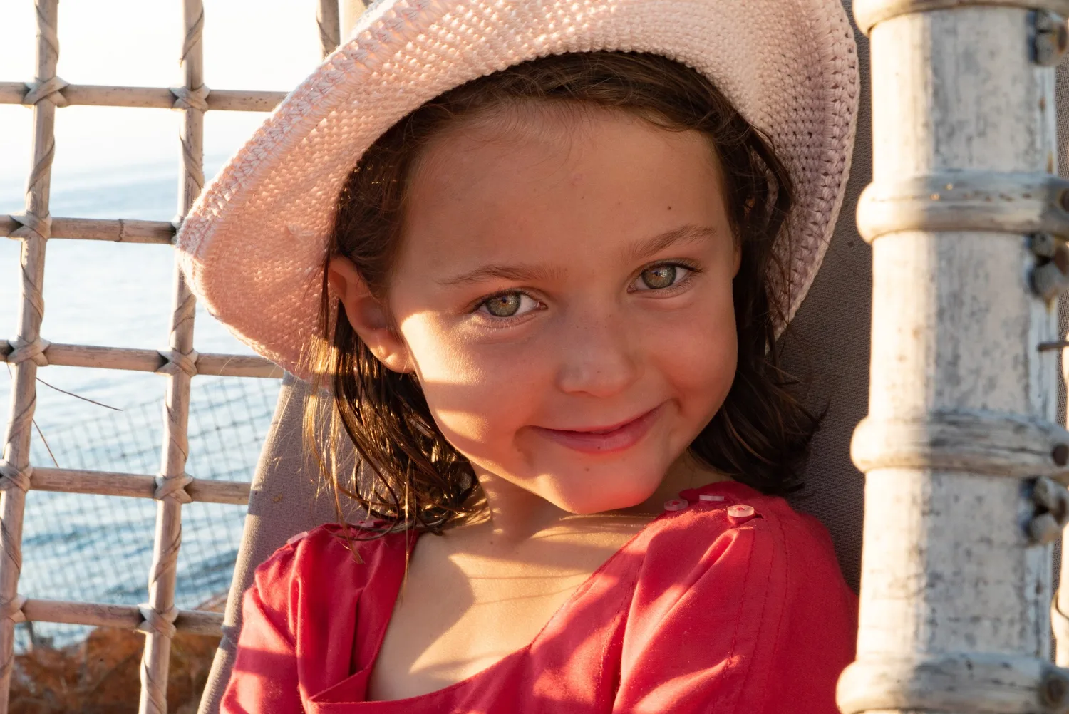 Petite fille en bonnet au crochet rose et haut rouge, assise dans un fauteuil de corde devant la mer, à Porquerolles.