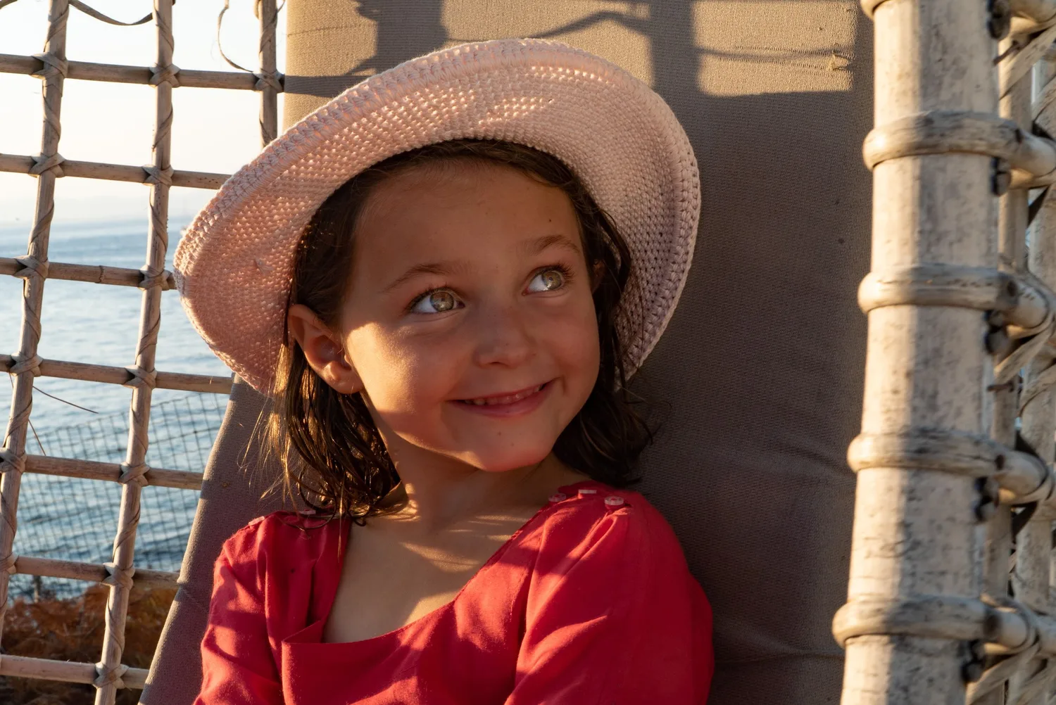 Enfant souriante en chapeau rose au crochet et blouse rouge près de la mer à Porquerolles.