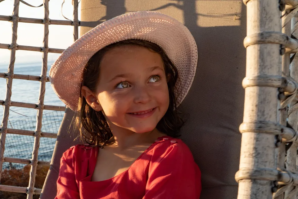 Enfant souriante en chapeau rose au crochet et blouse rouge près de la mer à Porquerolles.