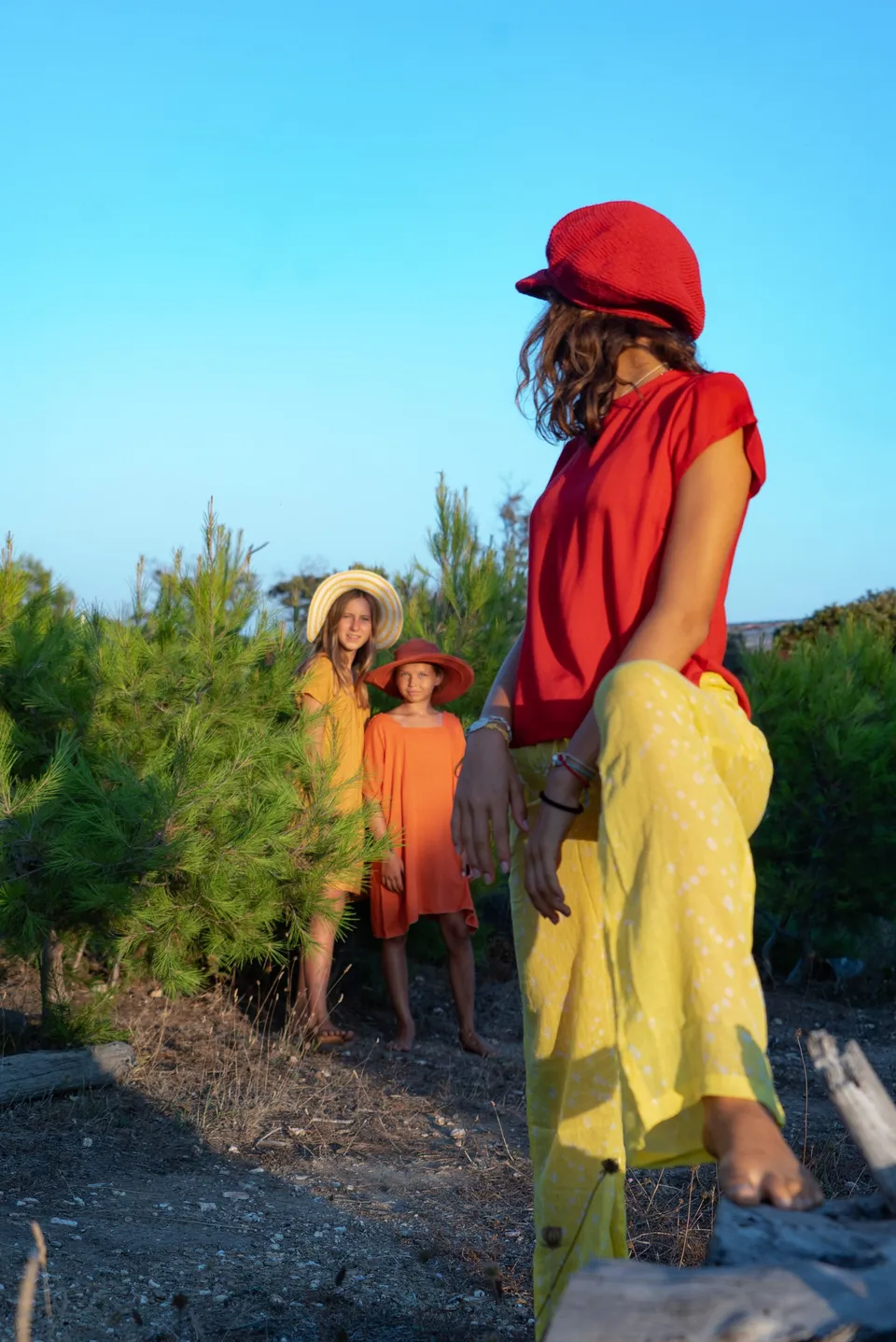 Femme et deux filles en rouge, jaune et orange, chapeaux colorés dans le maquis à l'heure dorée à Porquerolles.