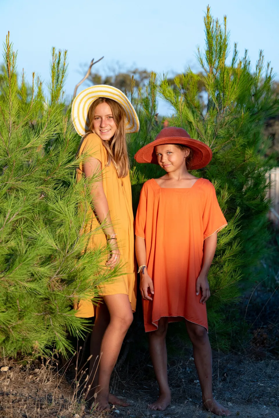 Deux filles en tuniques jaune et orange, chapeaux à larges bords, devant les pins du maquis à Porquerolles.