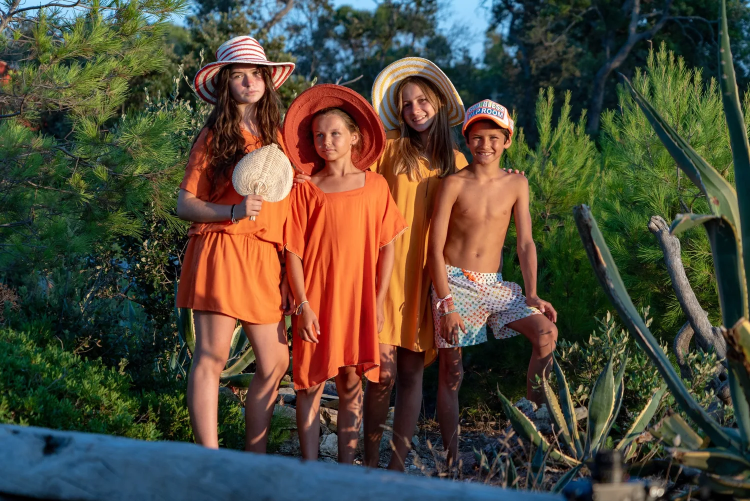Quatre enfants en tuniques orange et jaune, chapeaux de paille dans le maquis à l'heure dorée à Porquerolles.