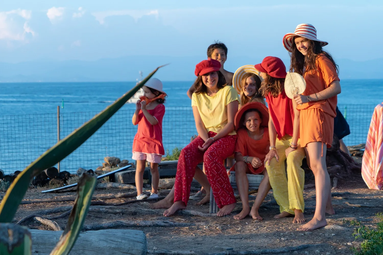 Enfants en tenues rouge jaune et orange, chapeaux de paille, terrasse face à la mer à Porquerolles.