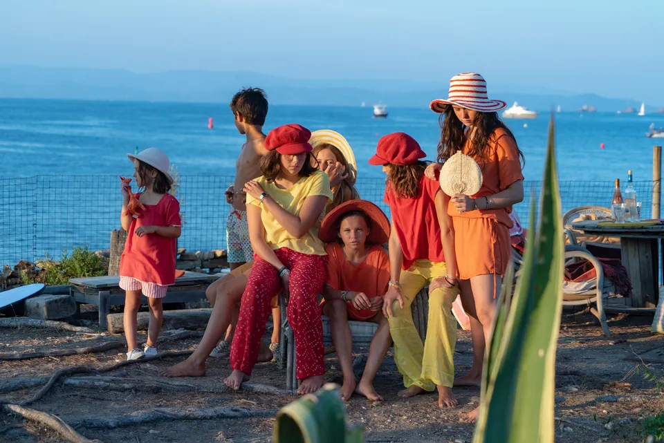 Groupe d'enfants en tuniques rouge et orange, chapeaux variés, posant face à la mer dorée à Porquerolles.