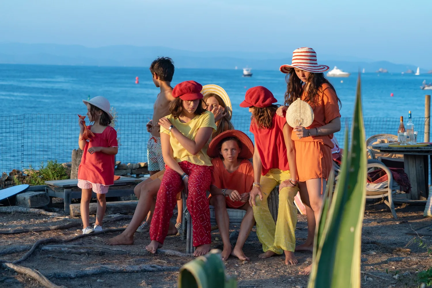 Groupe d'enfants en tuniques rouge et orange, chapeaux variés, posant face à la mer dorée à Porquerolles.