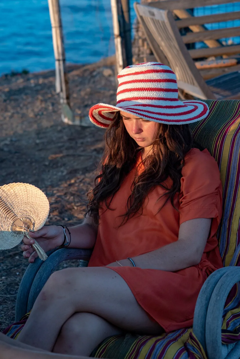 Jeune fille en robe orange et chapeau rayé, éventail de vannerie, assise face à la mer à Porquerolles.